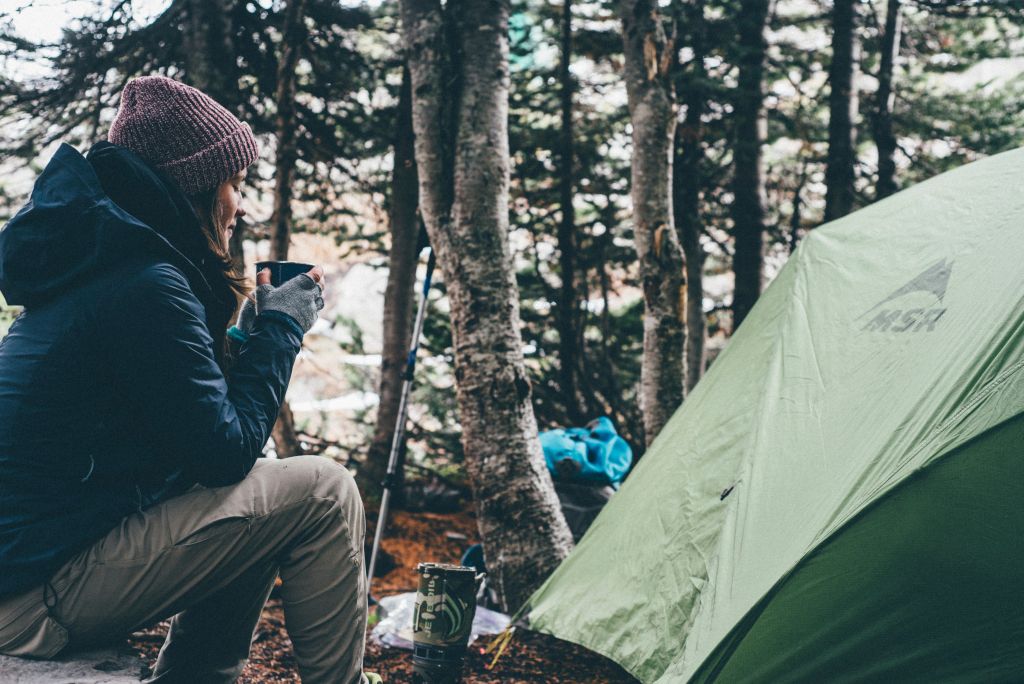 Woman sitting by a green tent in a forest, holding a mug.