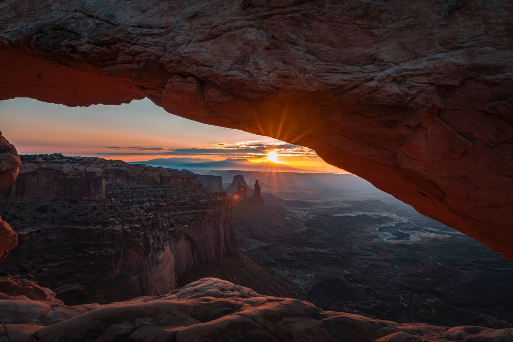 Sunrise seen through a stone arch with a canyon and distant rock formations in the background.