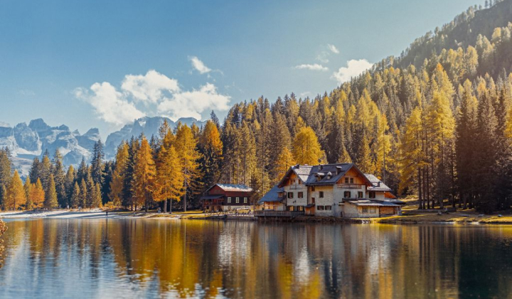 Lake surrounded by autumn trees and a mountain chalet reflected in the calm water. Snow-capped mountain peaks in the background.